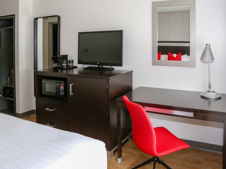 Une chambre d’hôtel avec un lit, un bureau avec un ordinateur, une chaise rouge, une télévision, un four à micro-ondes et une lampe.