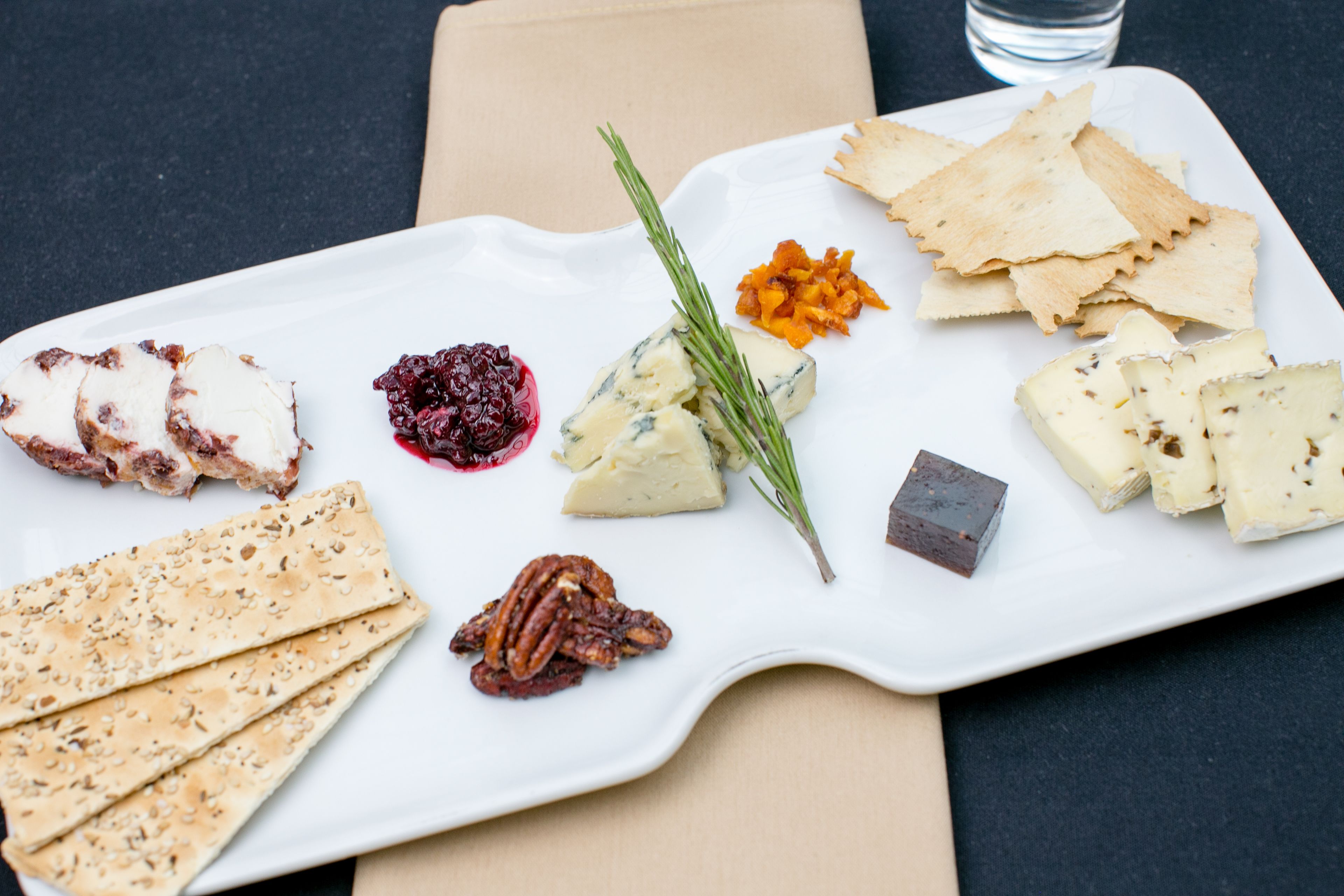 Un plato rectangular blanco con varios quesos, galletas y una ramita de romero.