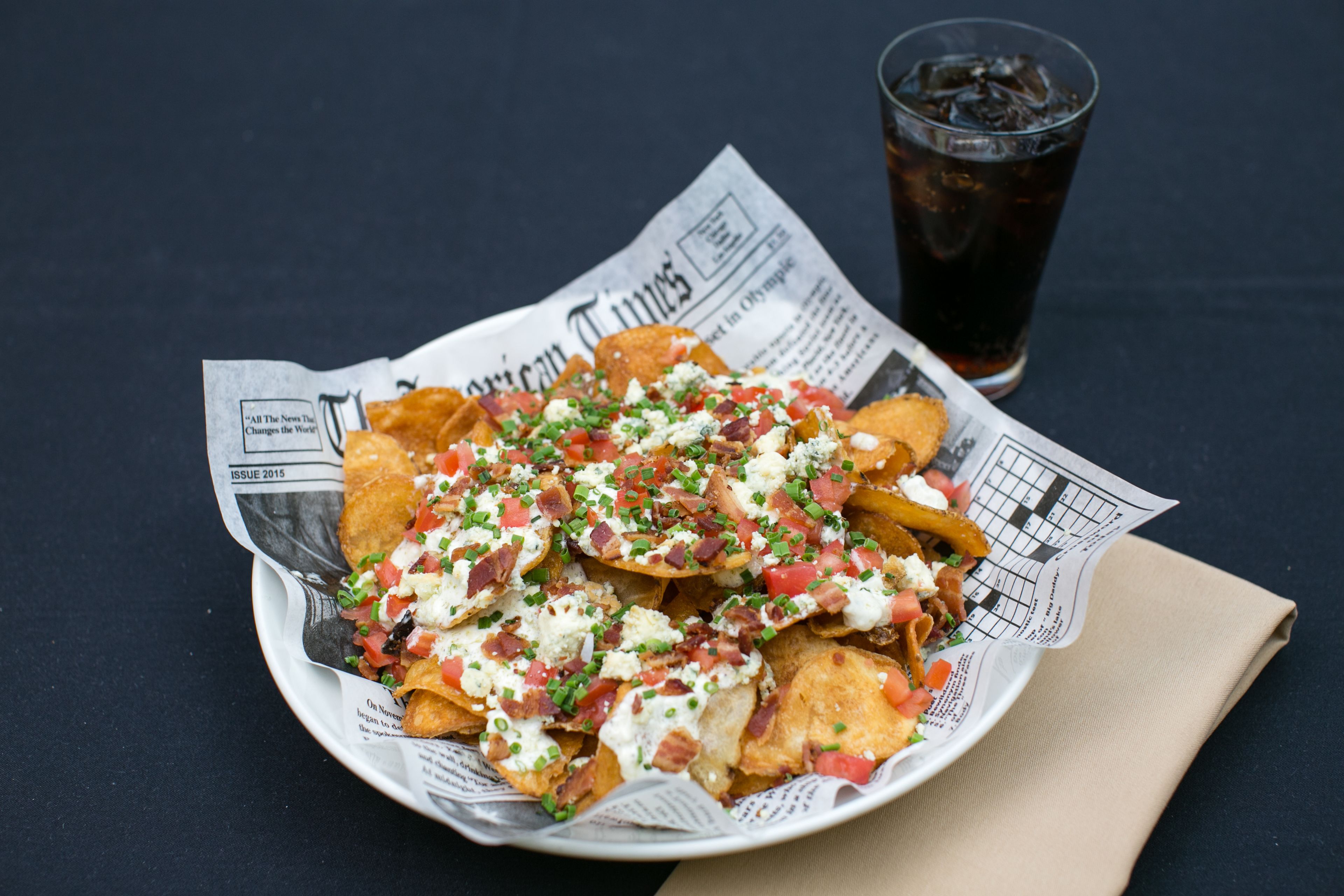 Nachos sobre un periódico, un vaso de refresco y una servilleta sobre una mesa.