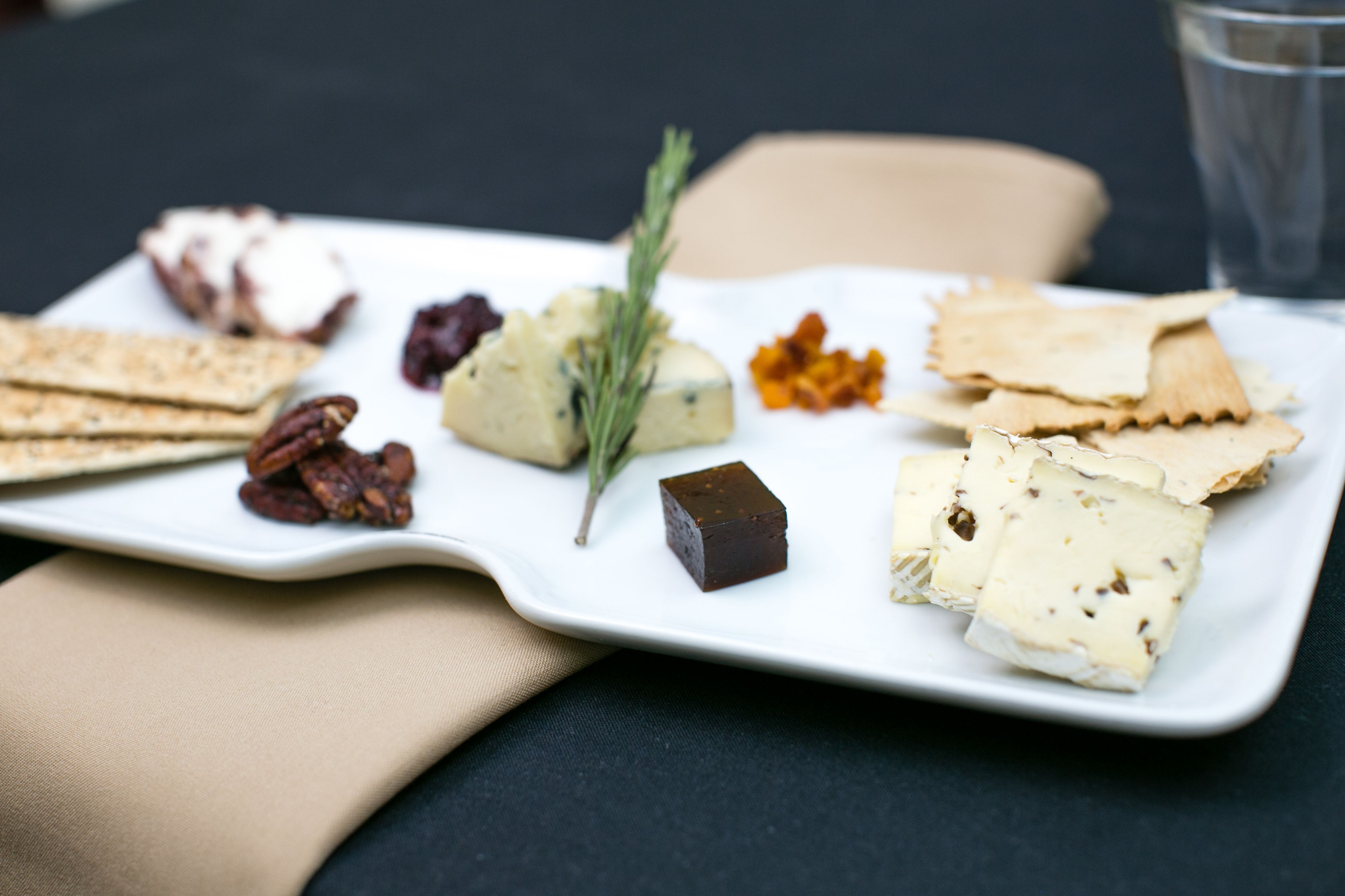 A white rectangular plate with various cheeses, crackers, and nuts on a dark tablecloth.