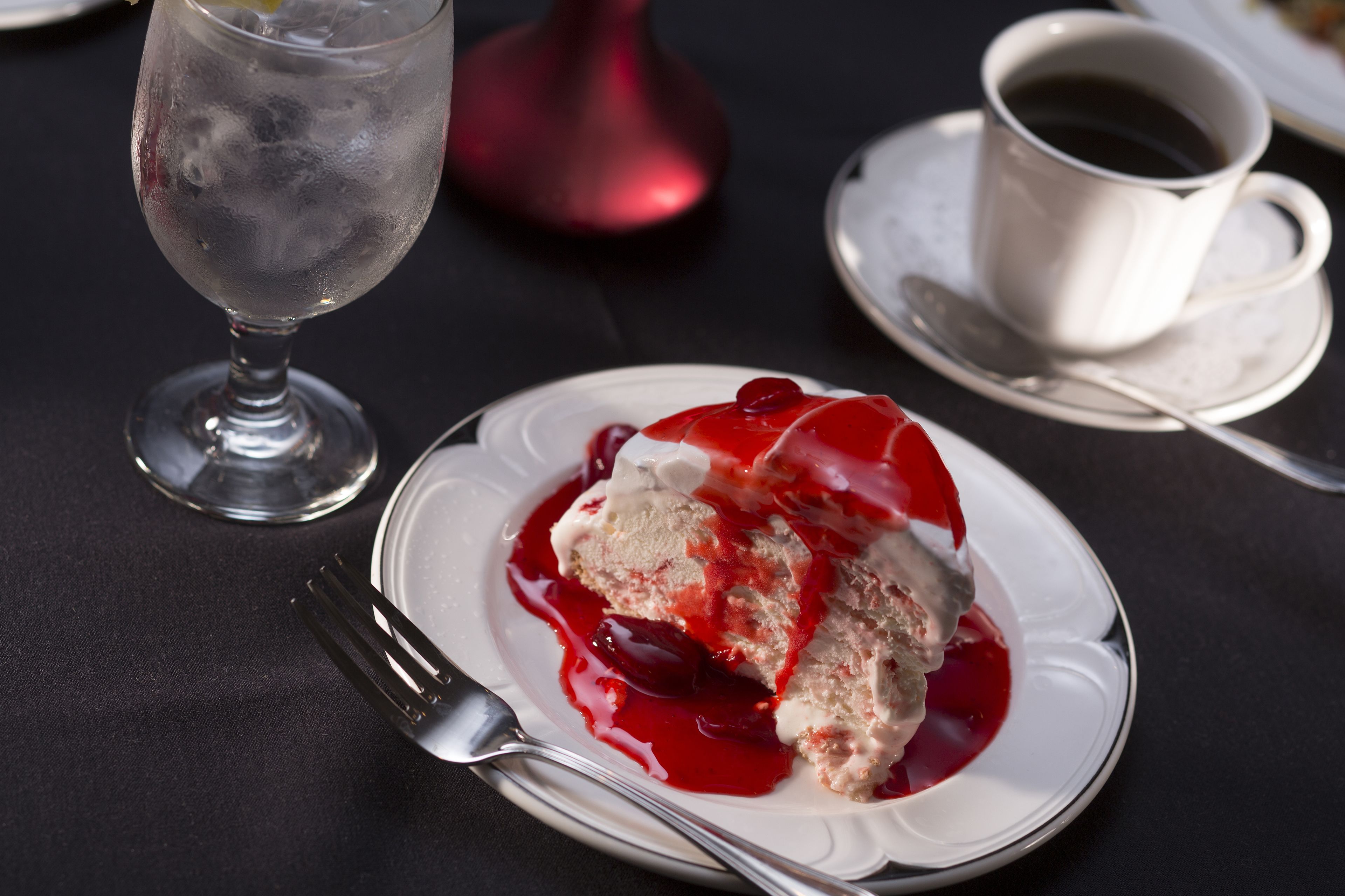 Un plato de postre con helado y salsa roja, un vaso de agua y una taza de café sobre una mesa.