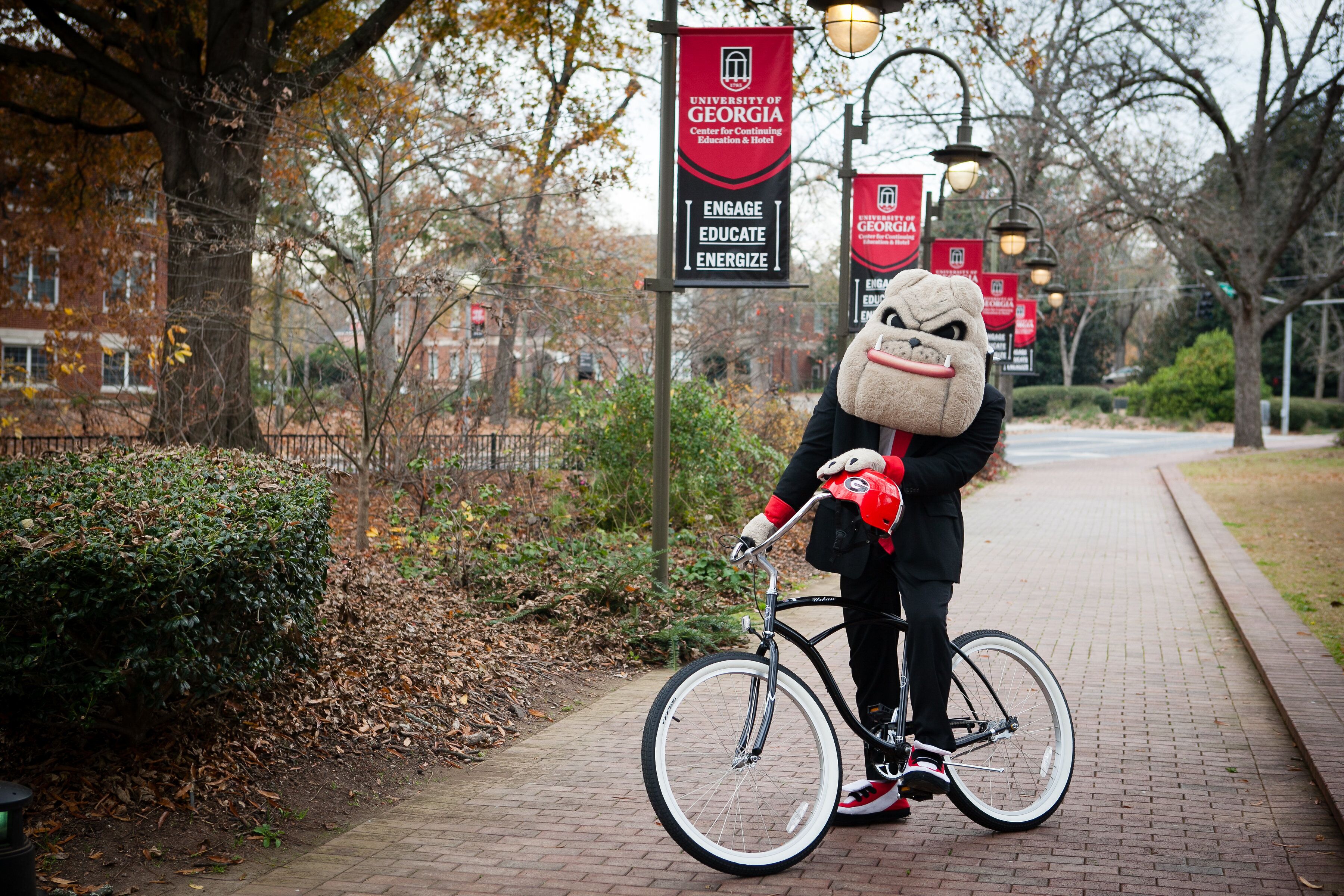 A mascot on a bicycle with a banner reading "University of Georgia" and "Engage Educate Energize" in the background.