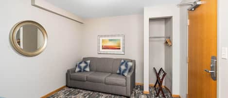 A room with a grey sofa, a round mirror, a framed painting, a wooden door, and a coat rack.