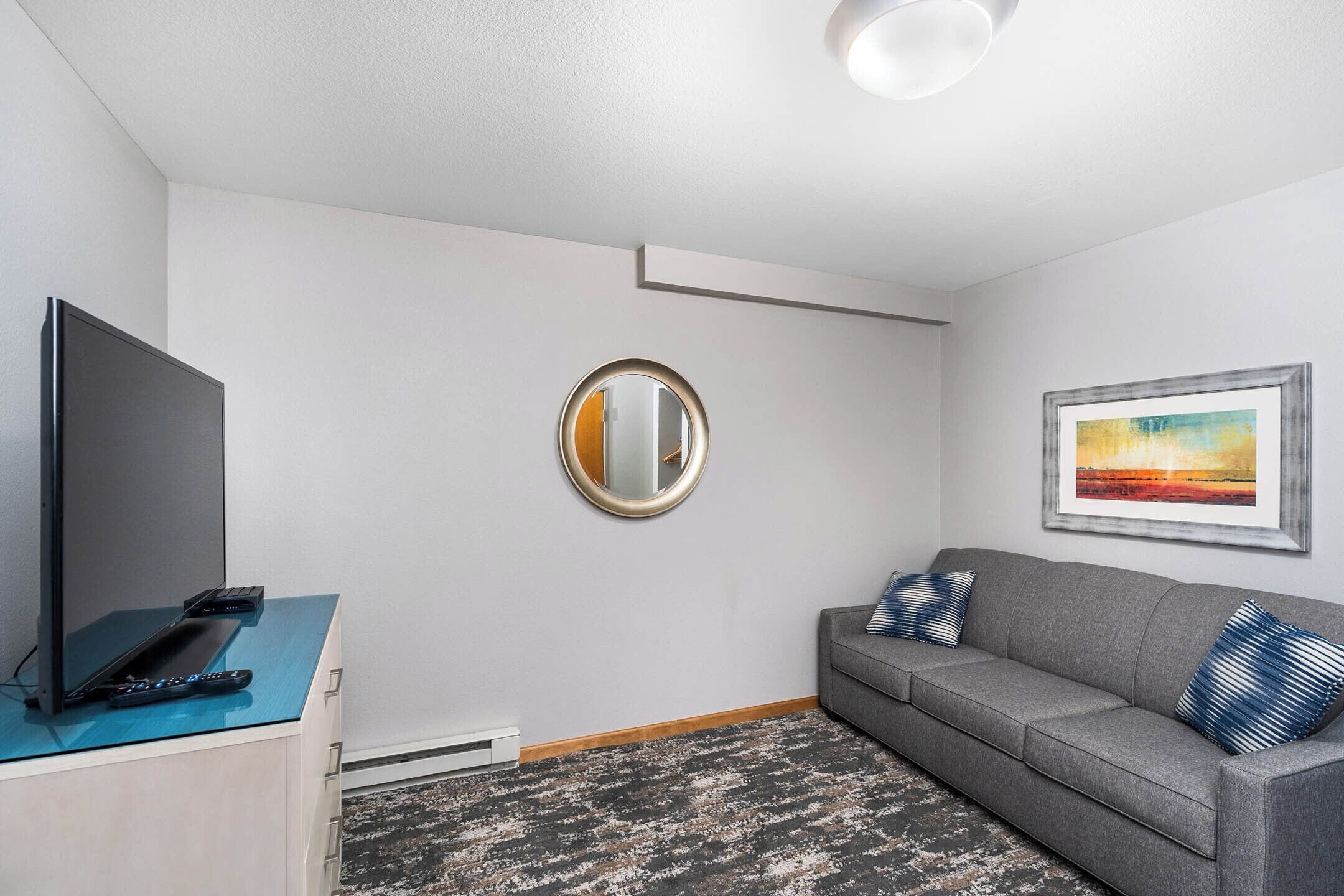 A modern living room with a flat-screen TV, a sofa with patterned cushions, a round mirror, and a framed painting on the wall.