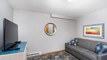 A modern living room with a flat-screen TV, a sofa with patterned cushions, a round mirror, and a framed painting on the wall.