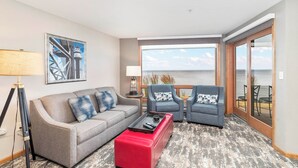 Habitación de hotel moderna con un sofá gris, dos sillones, un reposapiés rojo y un amplio ventanal con vistas al mar.