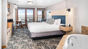 A hotel room with a large bed, a desk with a chair, a view of the sea, a bathroom with a bathtub, and a small kitchenette.