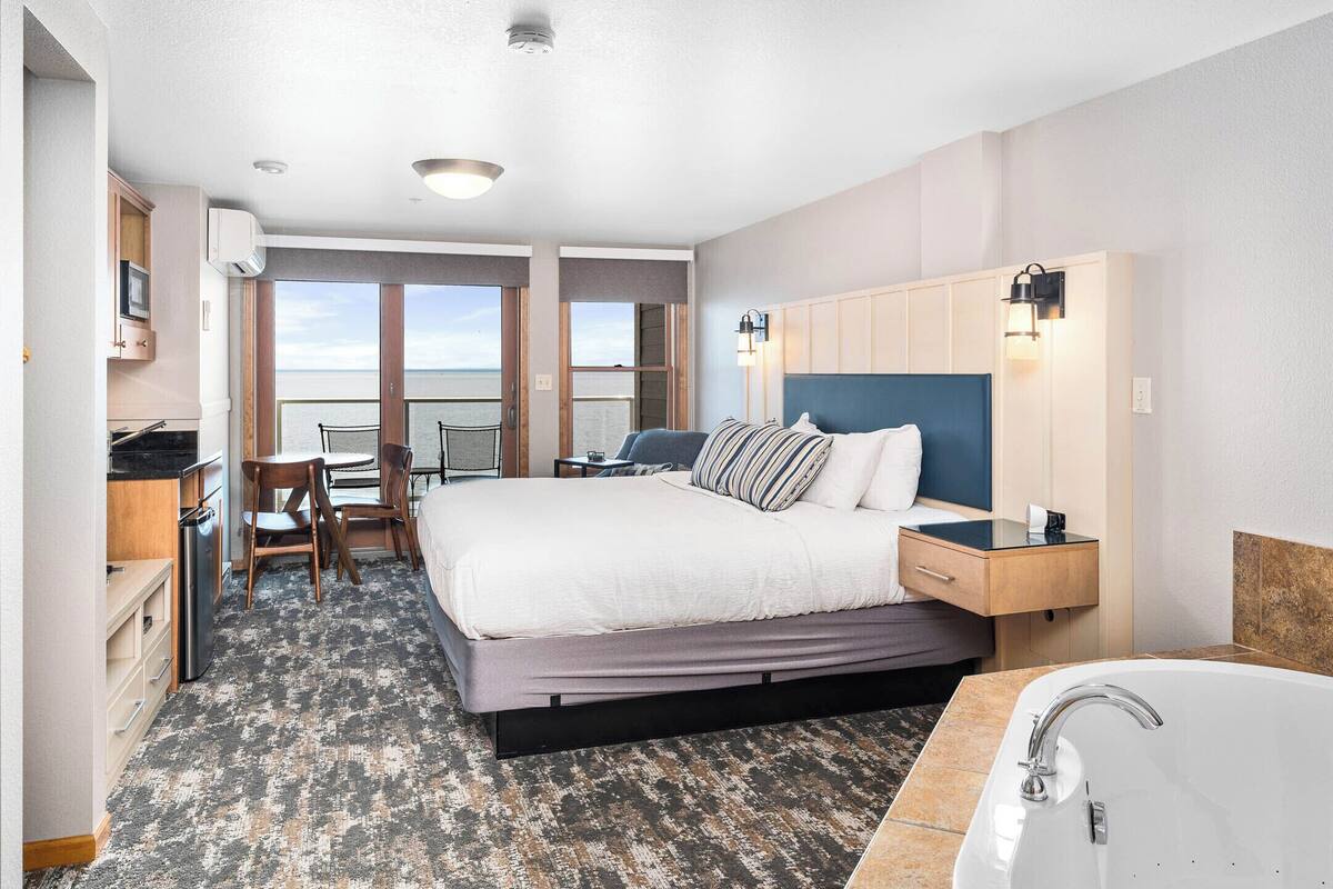 A hotel room with a large bed, a desk with a chair, a view of the sea, a bathroom with a bathtub, and a small kitchenette.