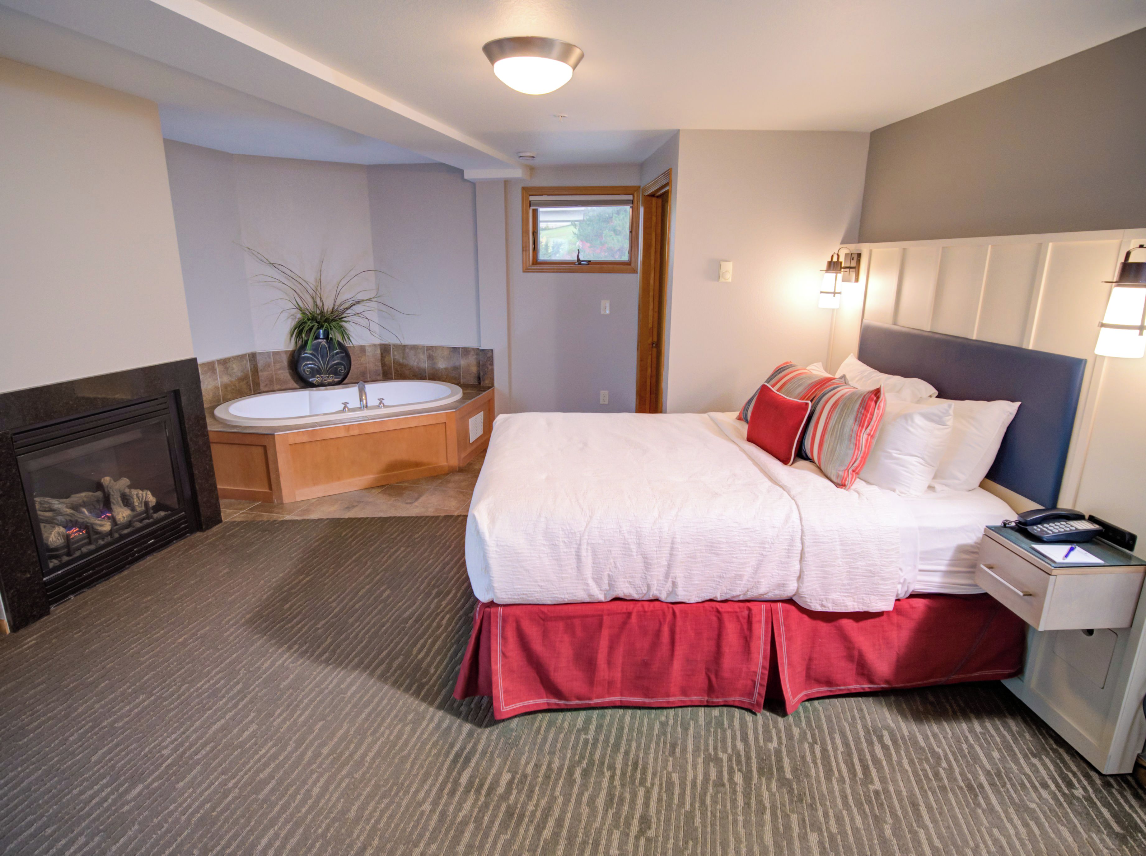 A bedroom with a large bed, a bedside table with a lamp, a nightstand, a fireplace, and a bathroom with a bathtub.