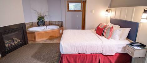 A bedroom with a large bed, a bedside table with a lamp, a nightstand, a fireplace, and a bathroom with a bathtub.