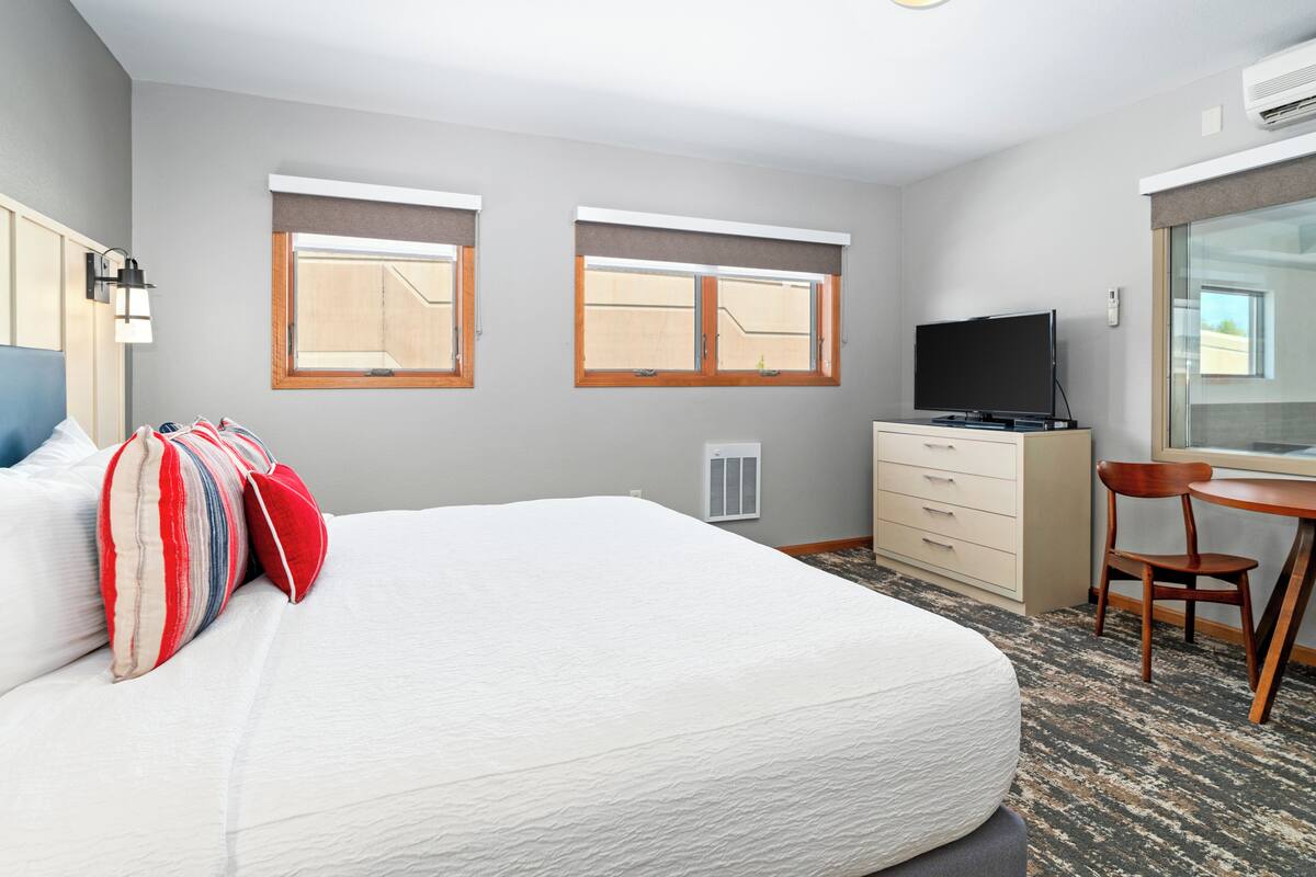 A hotel room with a large bed, two windows, a TV, a desk, and a chair.