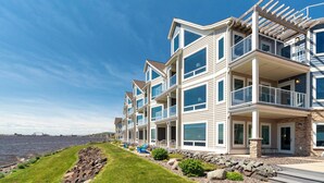A multi-story waterfront building with balconies, large windows, and a grassy area with rocks.