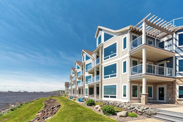 A multi-story waterfront building with balconies, large windows, and a grassy area with rocks. - Beacon Pointe on Lake Superior (Duluth)
