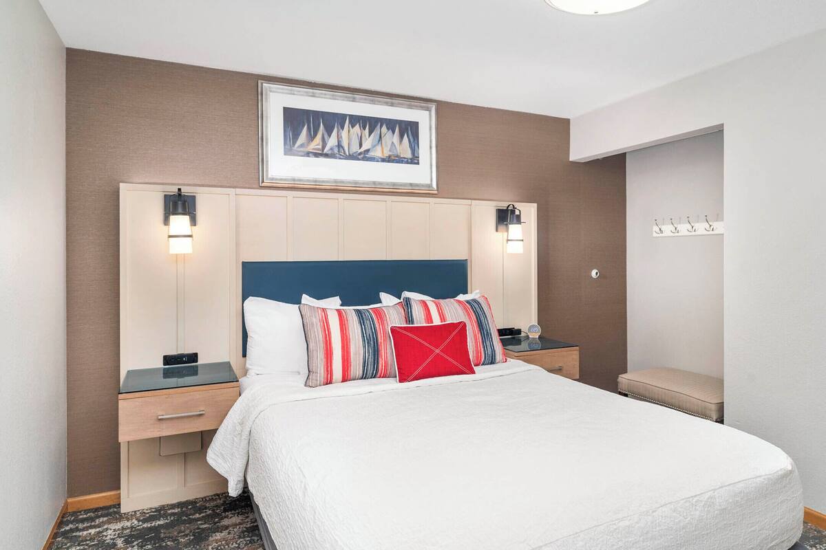 A hotel room with a large bed, two bedside tables, a nightstand, a framed picture on the wall, and a bench by the door.