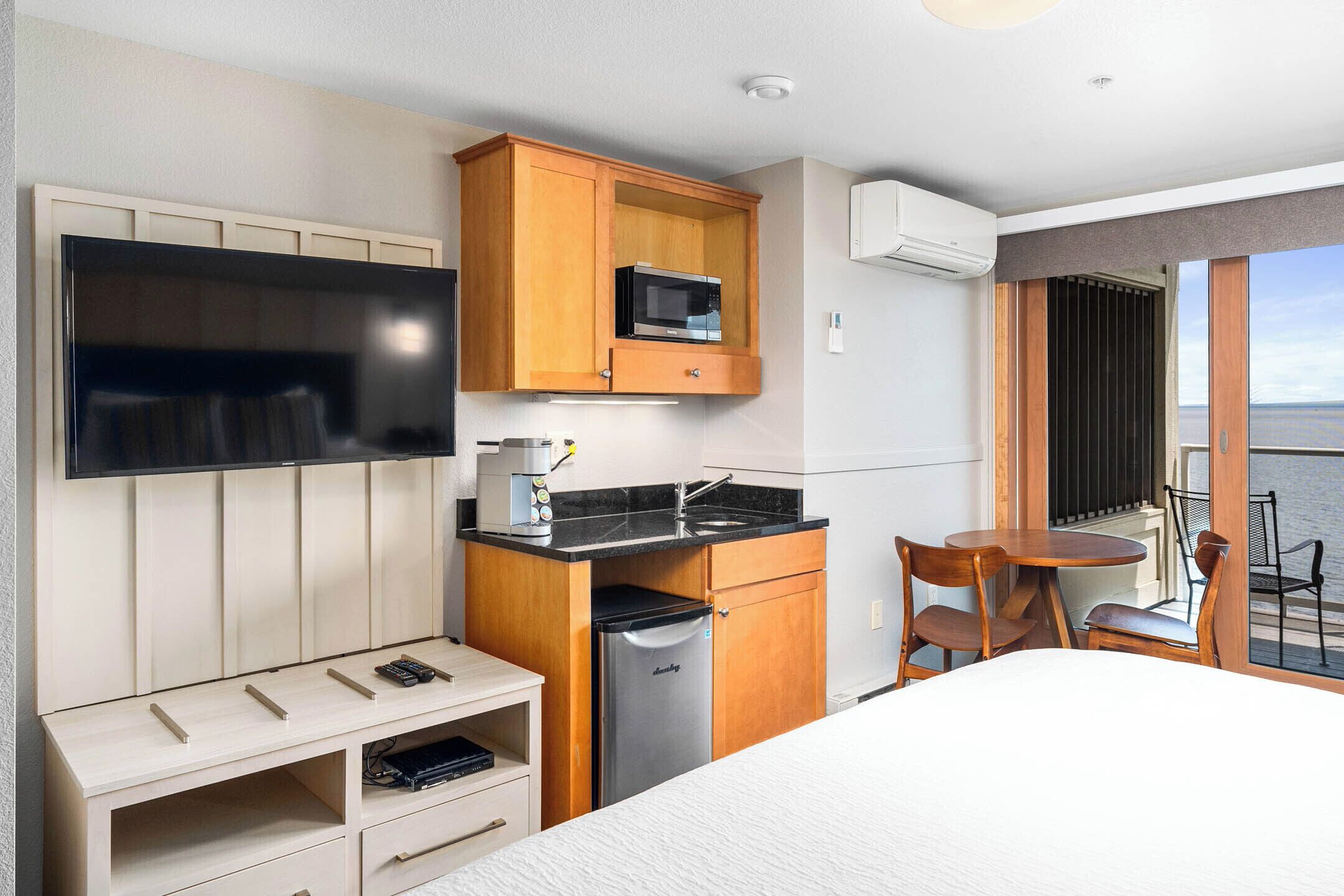 A compact hotel room with a bed, a TV, a kitchenette with a microwave, a small fridge, and a dining area with a table and chairs.