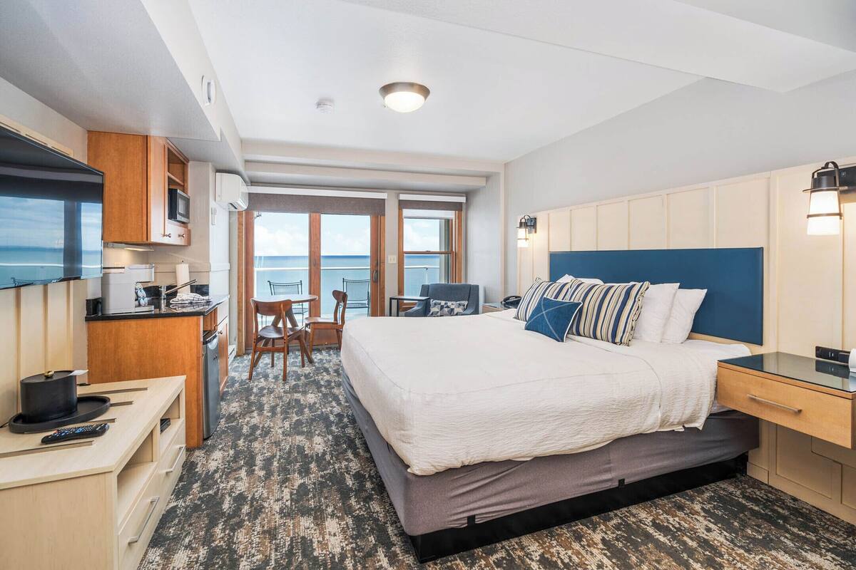 A modern hotel room with a large bed, a desk, a chair, a TV, and a view of the sea.