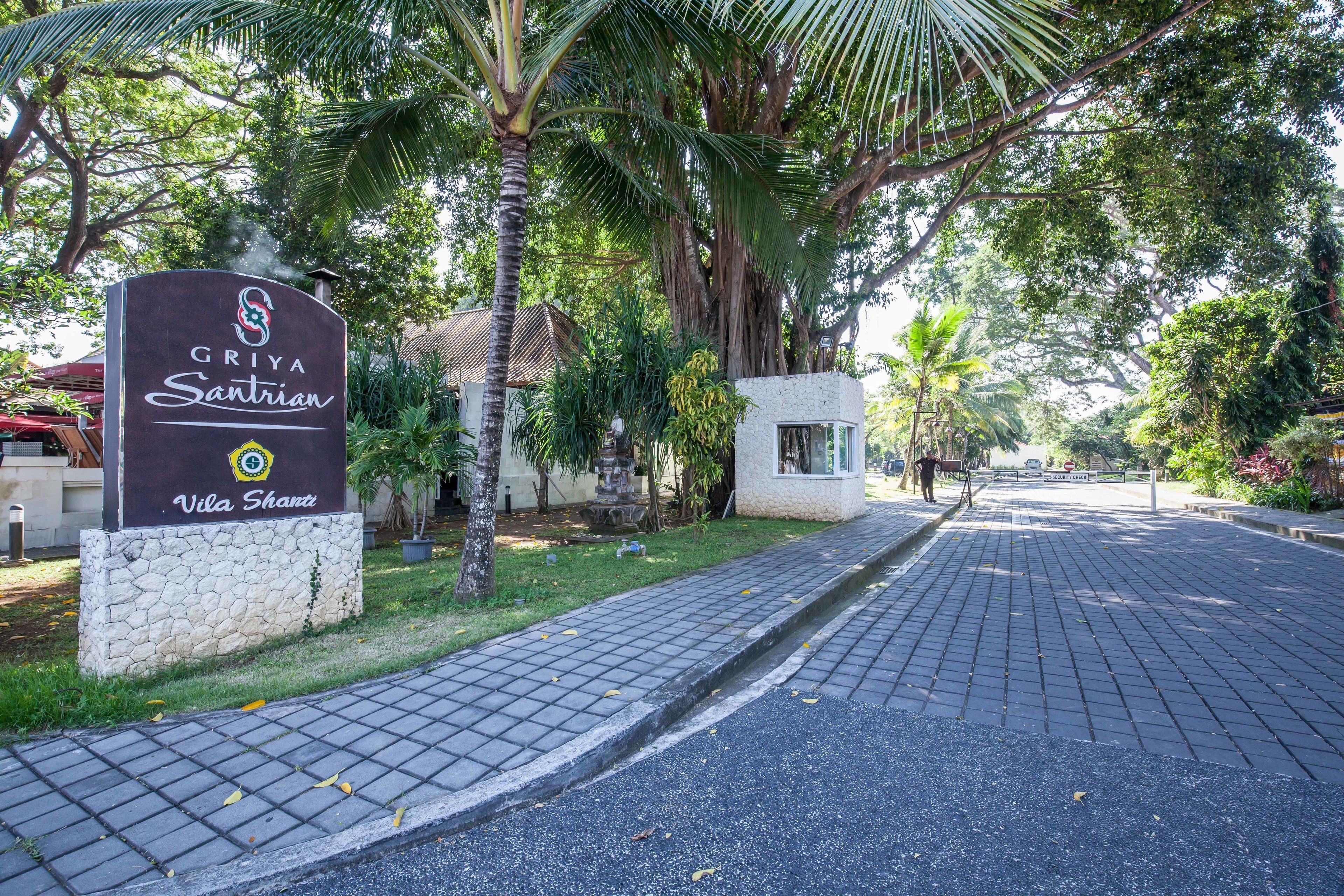 A sign for Griya Santian with Vila Sharzi, surrounded by lush greenery and a paved pathway.