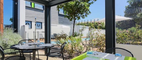 A modern outdoor seating area with a table set for two, a pool in the background, and a view of a building with large windows.