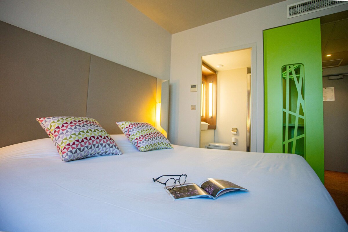 Een hotelkamer met een groot bed, twee kussens, een paar brillen en een open tijdschrift.
