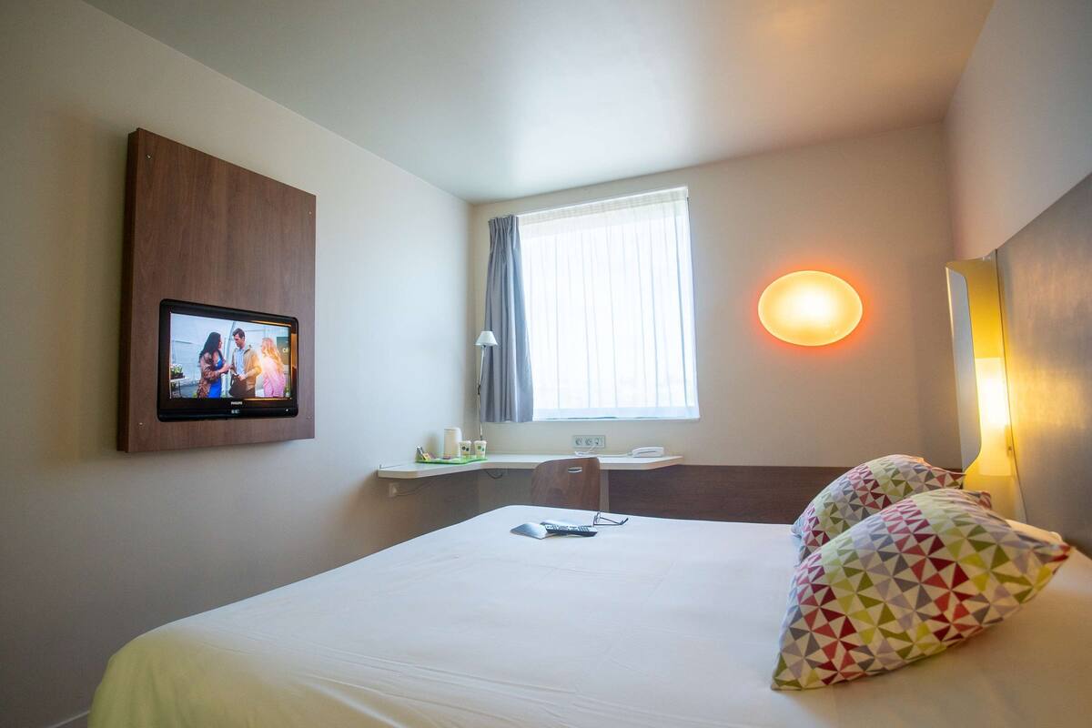 Hotelkamer met een bed, een aan de muur bevestigde televisie, een bureau met lamp en een raam met gordijnen.