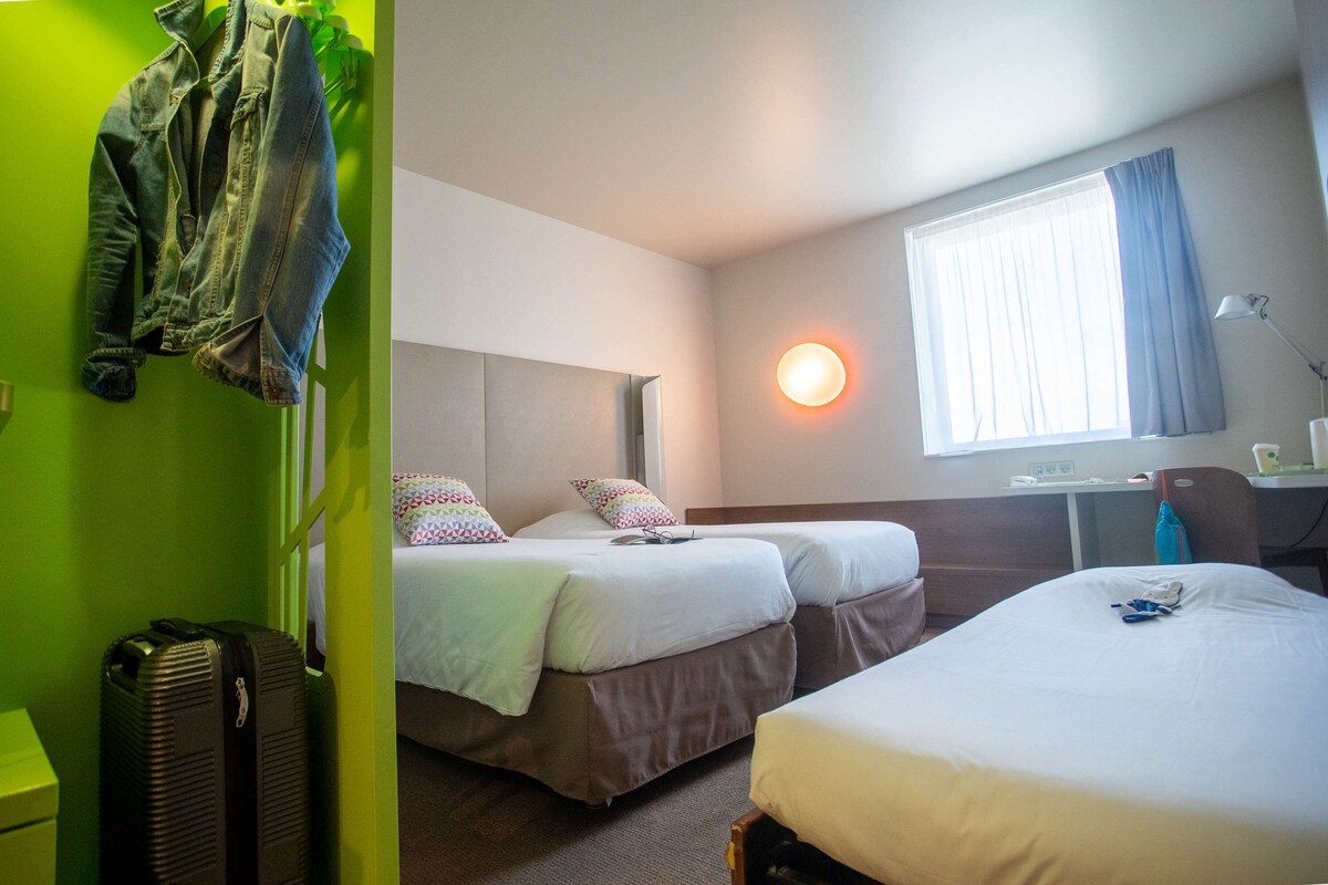Een hotelkamer met twee bedden, een bureau, een stoel, een lamp en een raam met gordijnen.