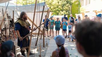 Une scène de reconstitution historique avec des participants vêtus de costumes d’époque, des structures en bois et une foule de spectateurs.