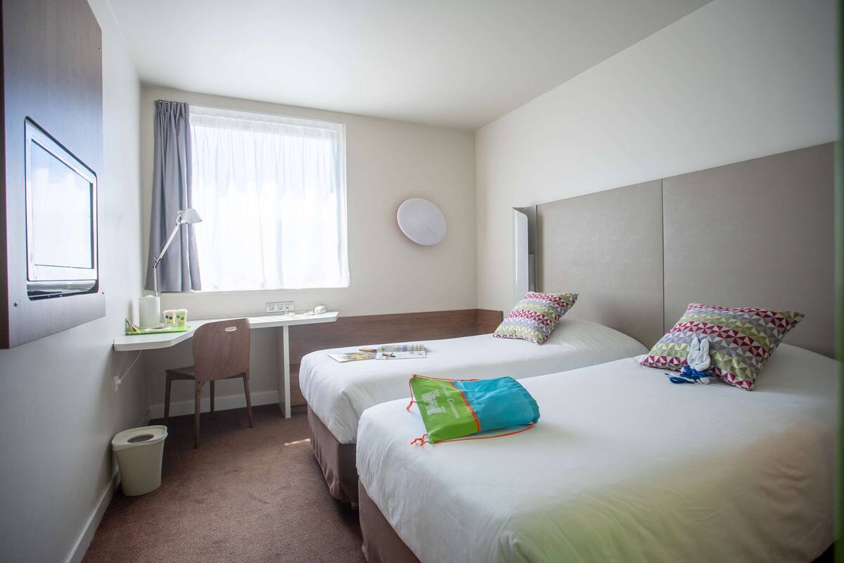 Hotelkamer met twee bedden, een bureau, een stoel, een prullenbak en een raam met gordijnen.
