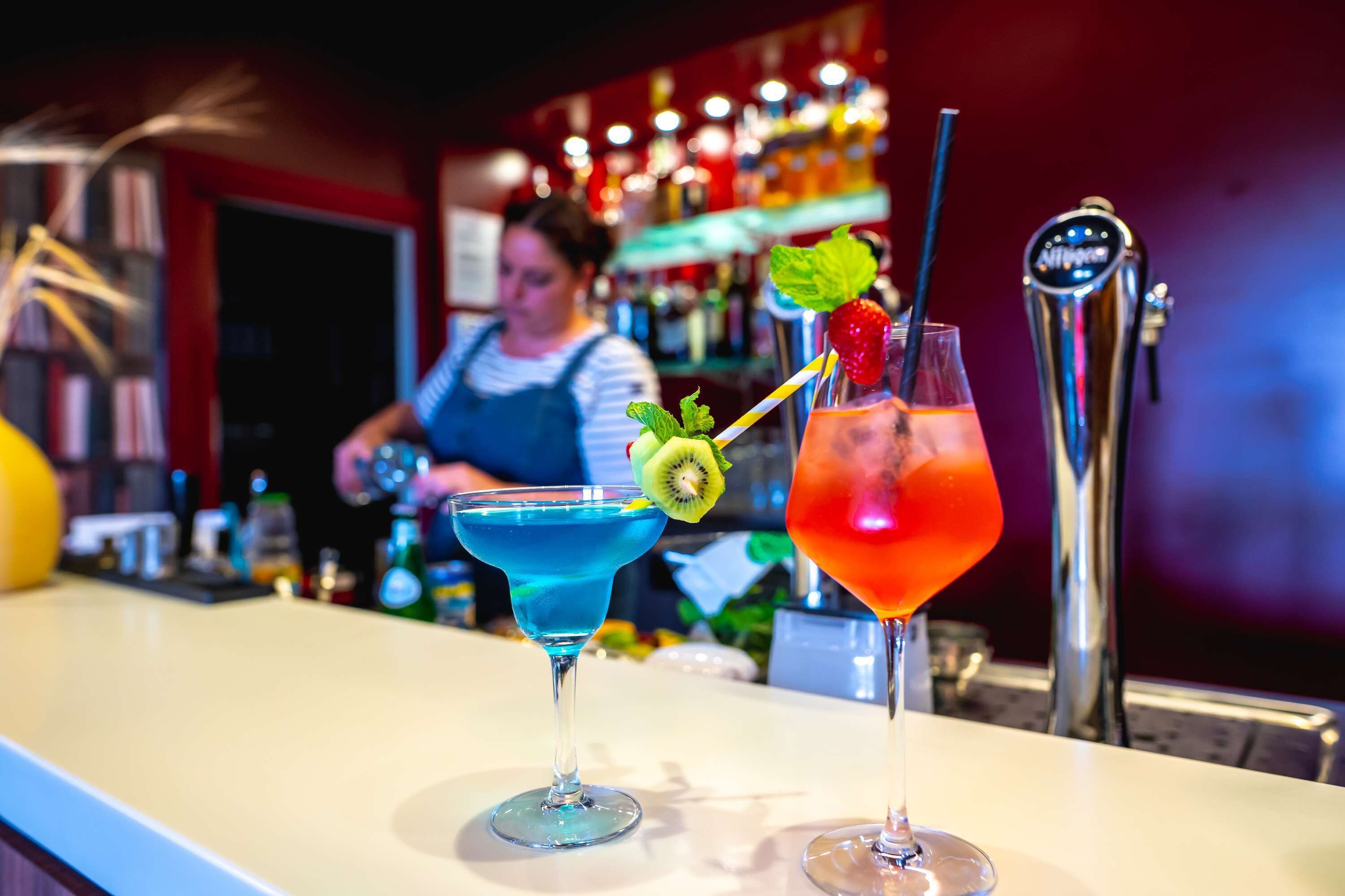 Two cocktails on a bar counter with a bartender in the background.