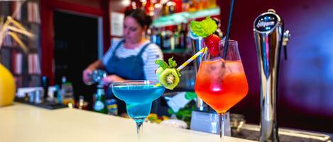Two cocktails on a bar counter with a bartender in the background.