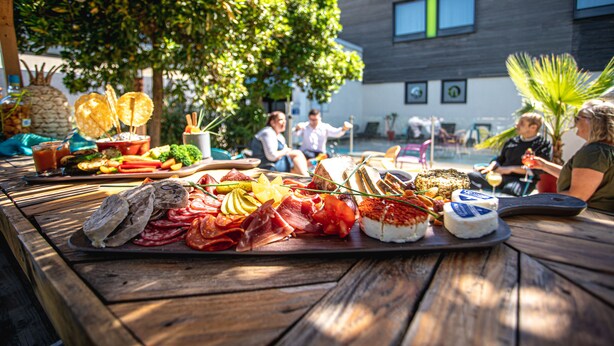 Un espace de restauration en plein air avec une table en bois dressée pour un repas, entouré de verdure et avec un bâtiment moderne en arrière-plan.
