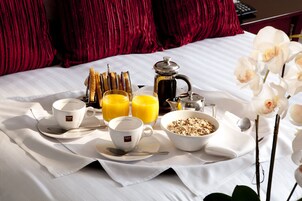 Un desayuno bien dispuesto sobre la cama, con un cabecero de terciopelo rojo, que incluye una cafetera, jugo de naranja, cereal y una taza con cuchara.