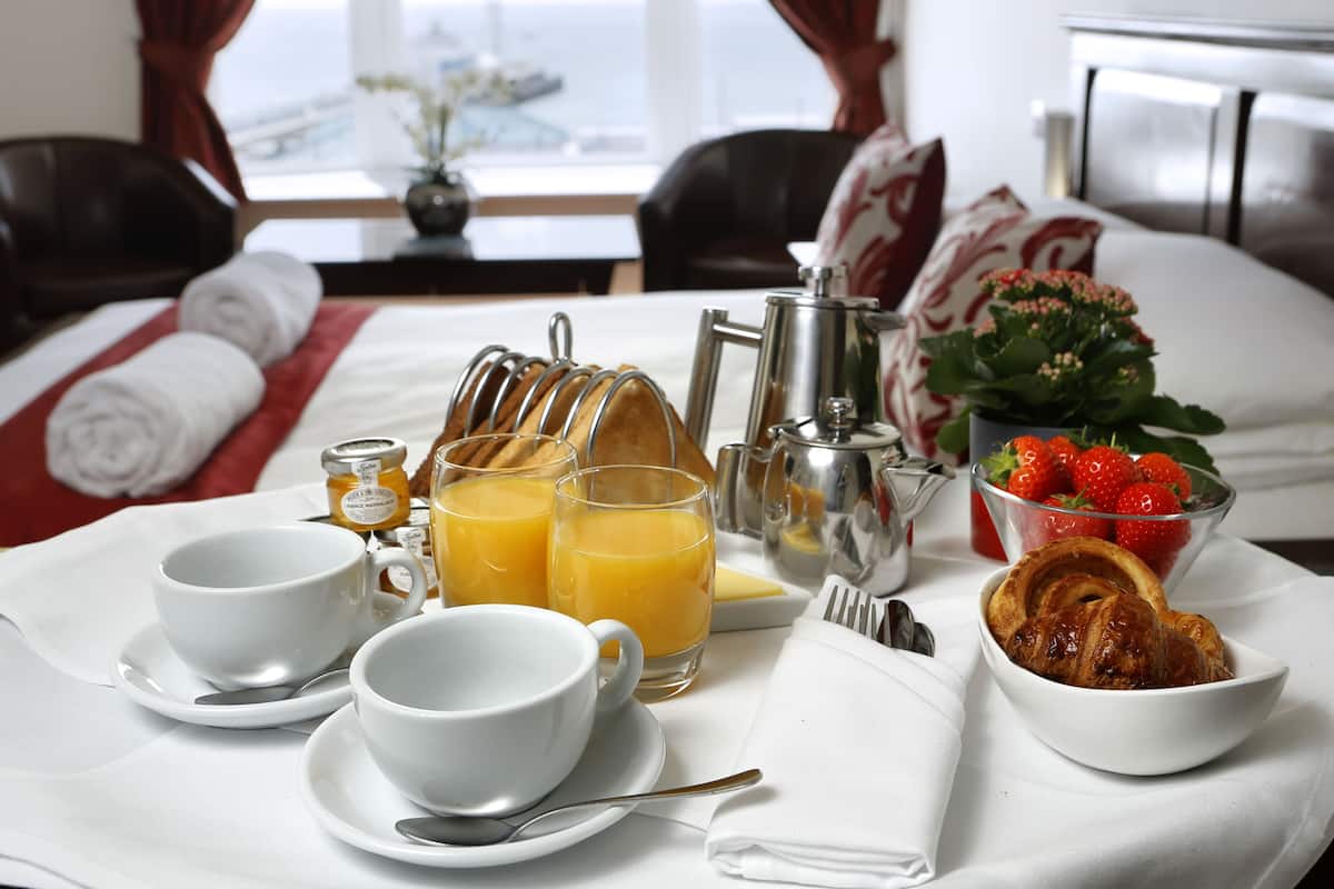 A breakfast setup with a bed, pillows, a vase with flowers, a fruit bowl, a coffee pot, and a bottle of honey.