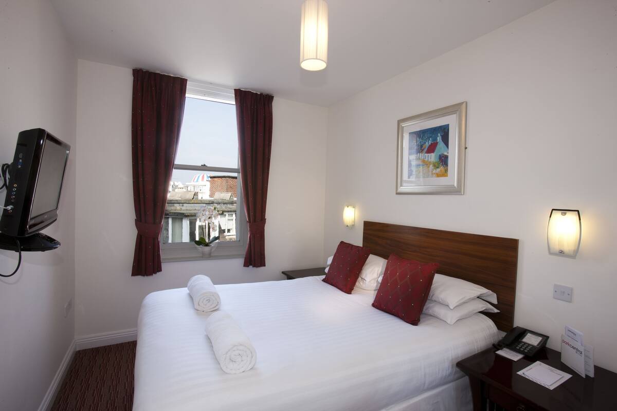 A hotel room with a neatly made bed, a bedside table with a phone and papers, a wall-mounted TV, a window with curtains, and a framed picture on the wall.