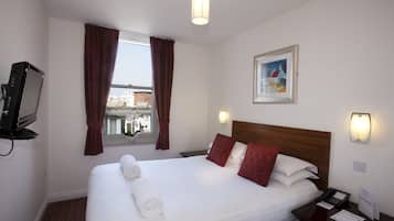 A hotel room with a neatly made bed, a bedside table with a phone and papers, a wall-mounted TV, a window with curtains, and a framed picture on the wall.