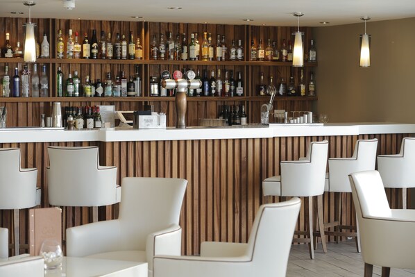 Un espace bar avec un mur en bois, des étagères remplies de bouteilles d’alcool et des tabourets de bar blancs.