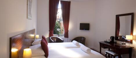 A hotel room with a bed, bedside lamps, a desk, a chair, a mirror, and a view of the outside through a window with curtains.