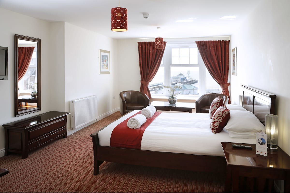 A hotel room with a large bed, two chairs, a table, a mirror, and a view of a cityscape.