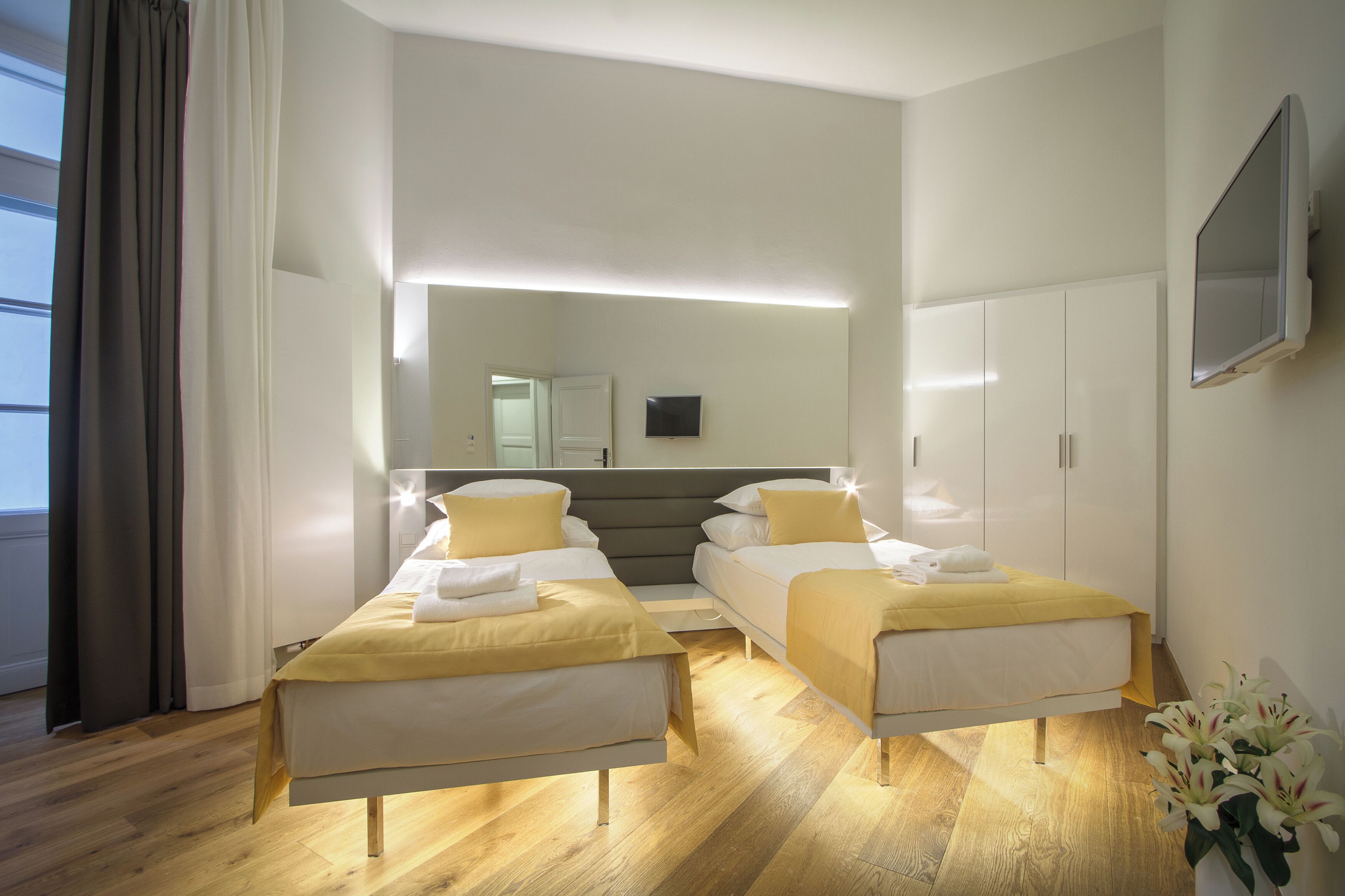 A hotel room with two single beds, a large mirror, a flat-screen TV, and a wooden floor.
