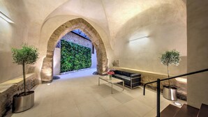 Un vestíbulo amplio con techo alto, arcos de piedra y una pared verde. Se observan dos plantas en macetas, un banco y una escalera.