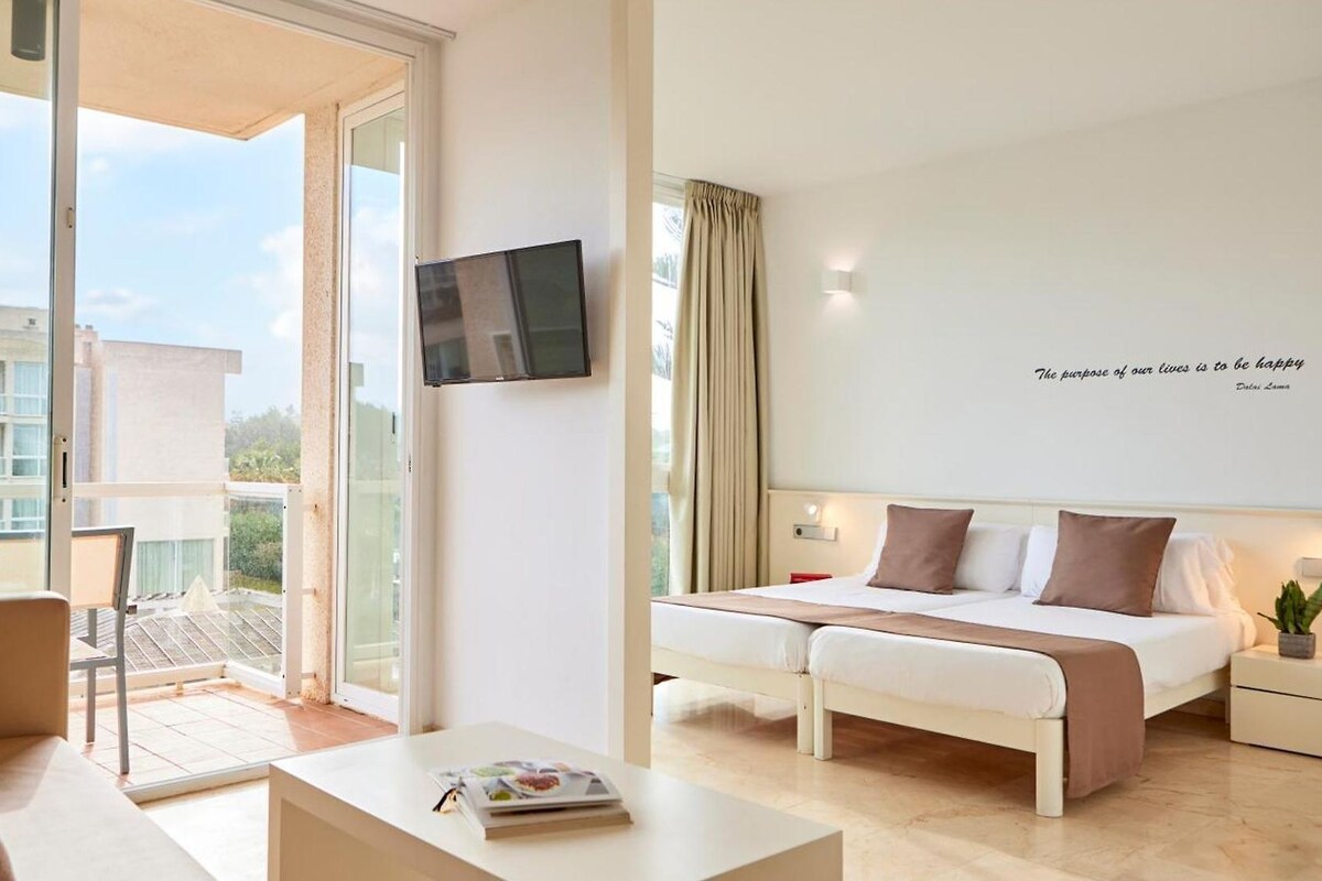 Une chambre d’hôtel moderne équipée d’un lit, d’une télévision, d’un balcon avec une table et des chaises, et offrant une vue sur les bâtiments environnants.