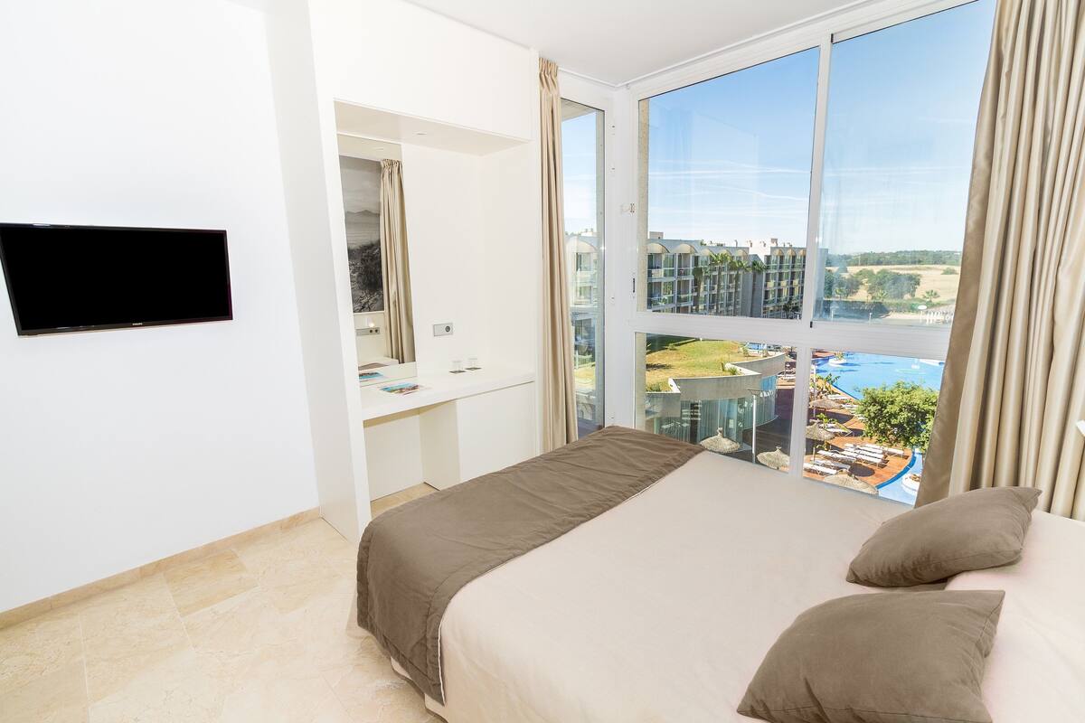 Habitación de hotel con cama, televisión, escritorio y un gran ventanal con vistas a una piscina y edificios.