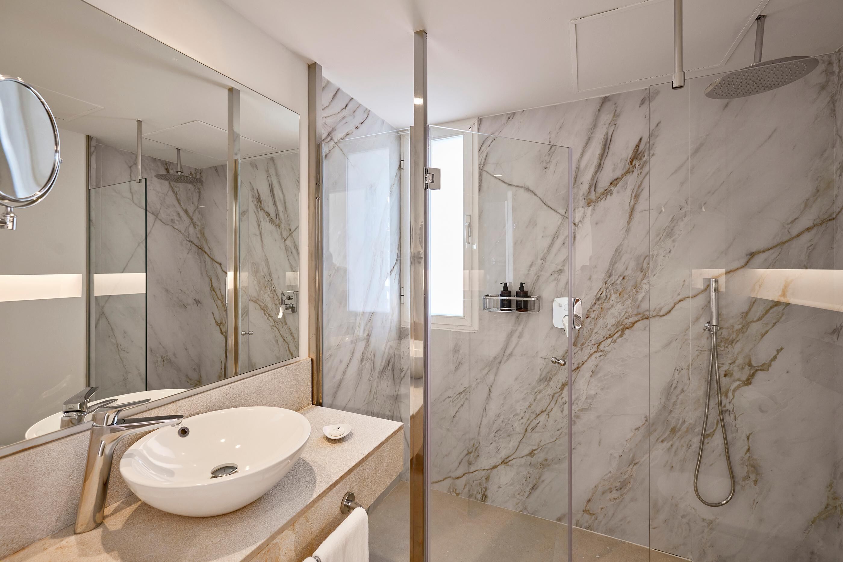 A modern bathroom with a marble vanity, a large mirror, a glass-enclosed shower, and a marble-tiled wall.