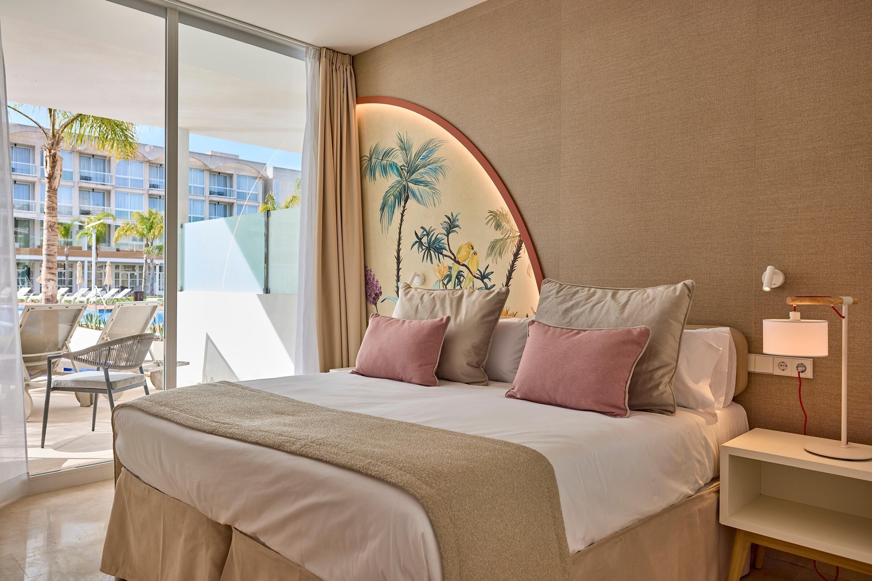 Habitación de hotel con una cama grande, una mesita de noche con lámpara, vista al área de la piscina y un mural en la pared.