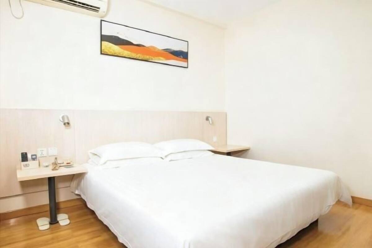 Une chambre d’hôtel avec un lit, une table de chevet avec un téléphone et une lampe, un climatiseur fixé au mur et un tableau au-dessus du lit.