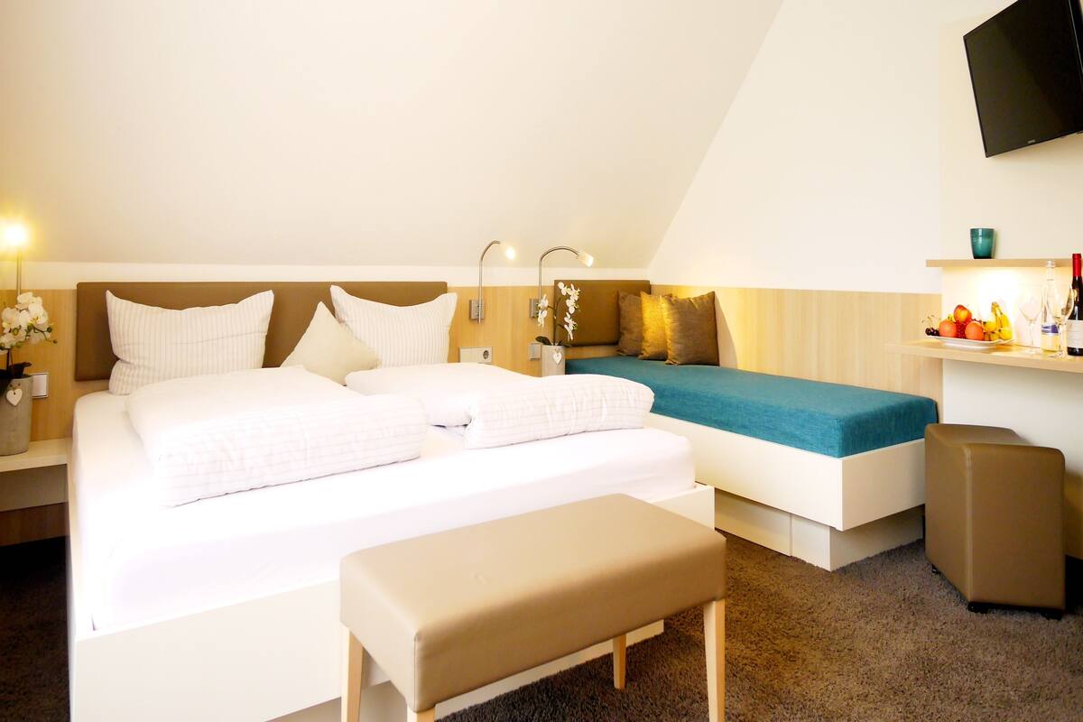 Hotelkamer met een groot bed, een kleiner bed, een bankje, een televisie en een plank met fruit en flessen.