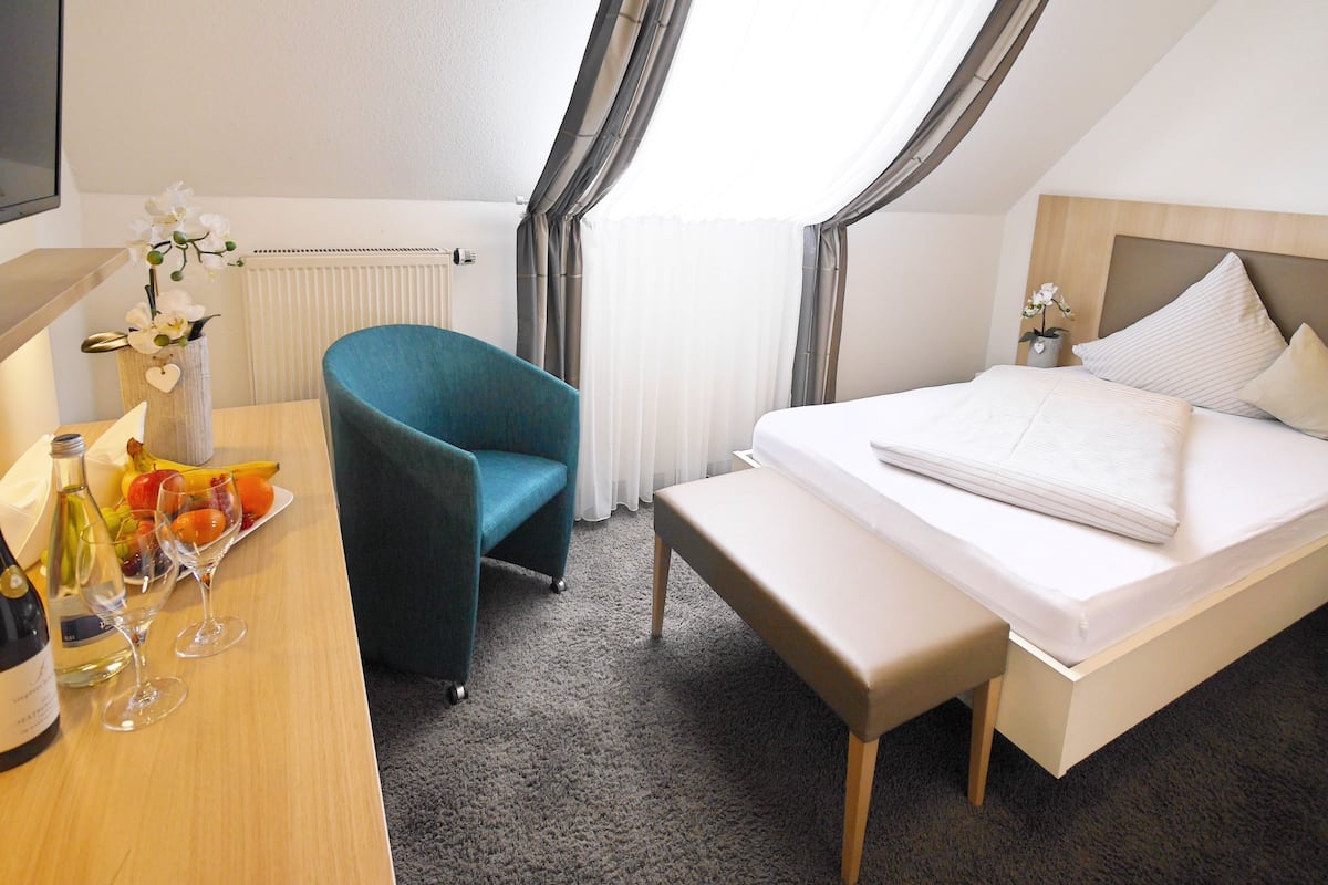 Hotelkamer met een bed, een bureau, een stoel, een bankje, een raam met gordijnen, en een tafeltje met fruit en een fles.