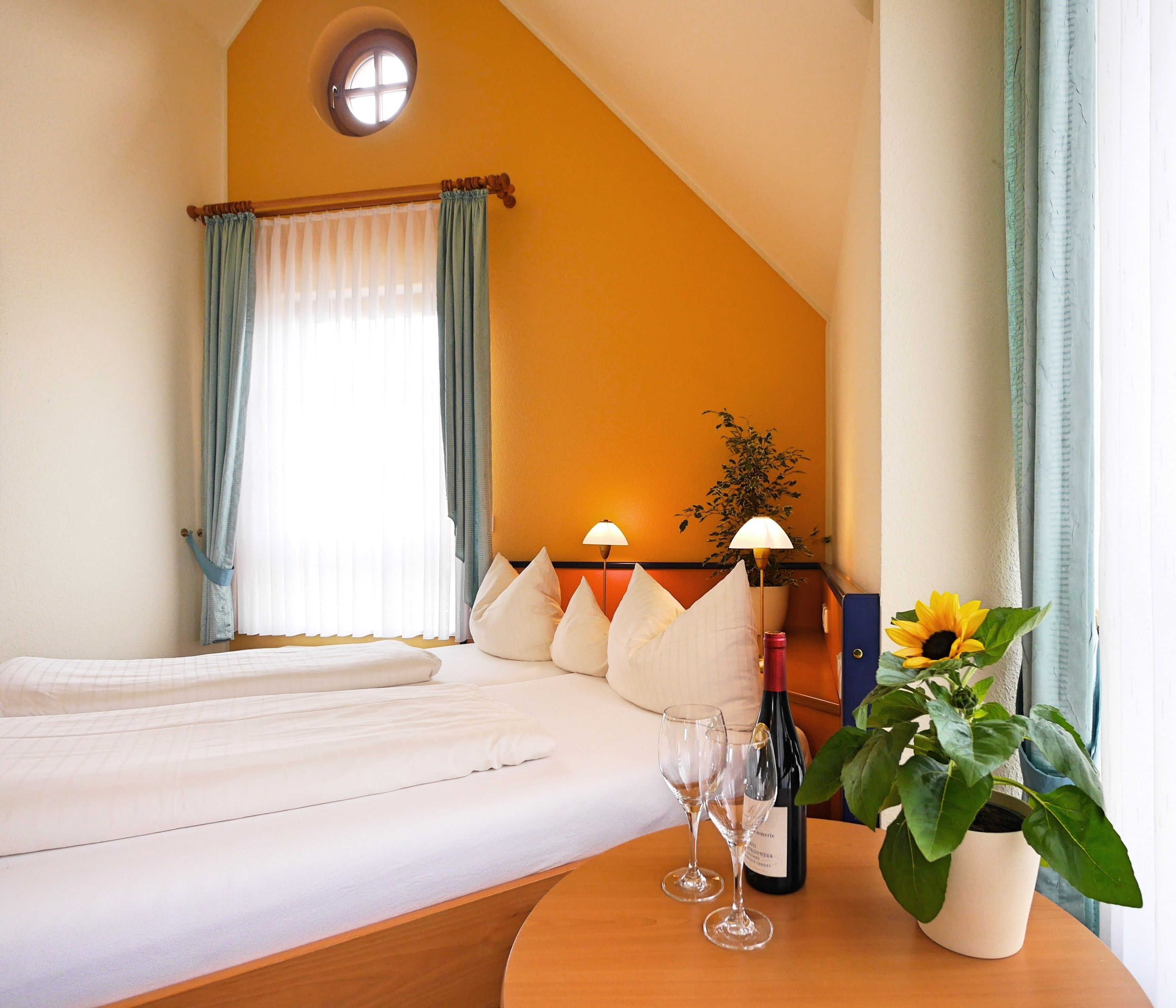 A hotel room with a bed, a table with wine glasses and a bottle, a potted plant, and a window with curtains.