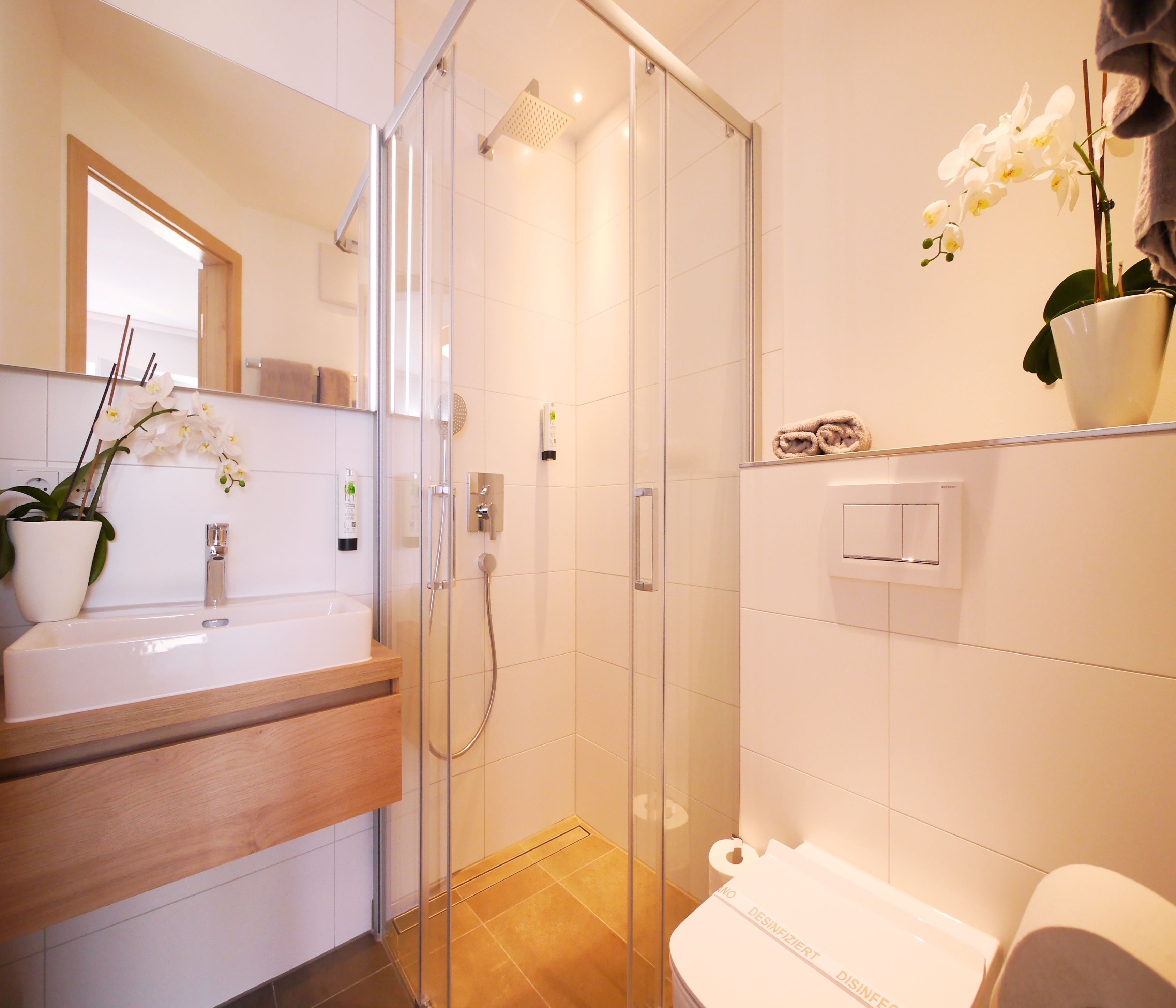 Un baño moderno con ducha envuelta en vidrio, lavamanos blanco y un inodoro con dispensador de toallas de papel.