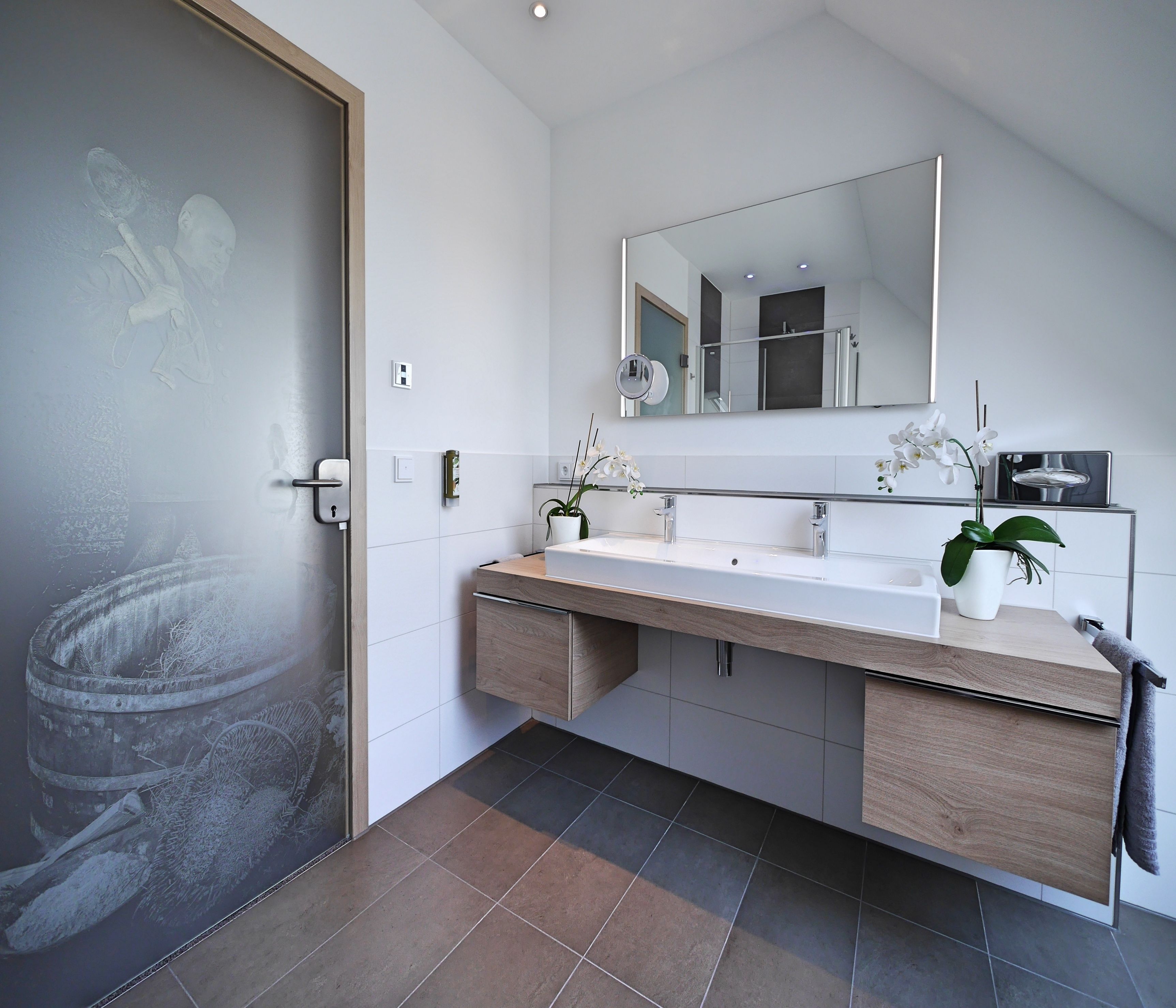 A modern bathroom with a large mirror, two sinks, wooden cabinets, and a potted orchid.