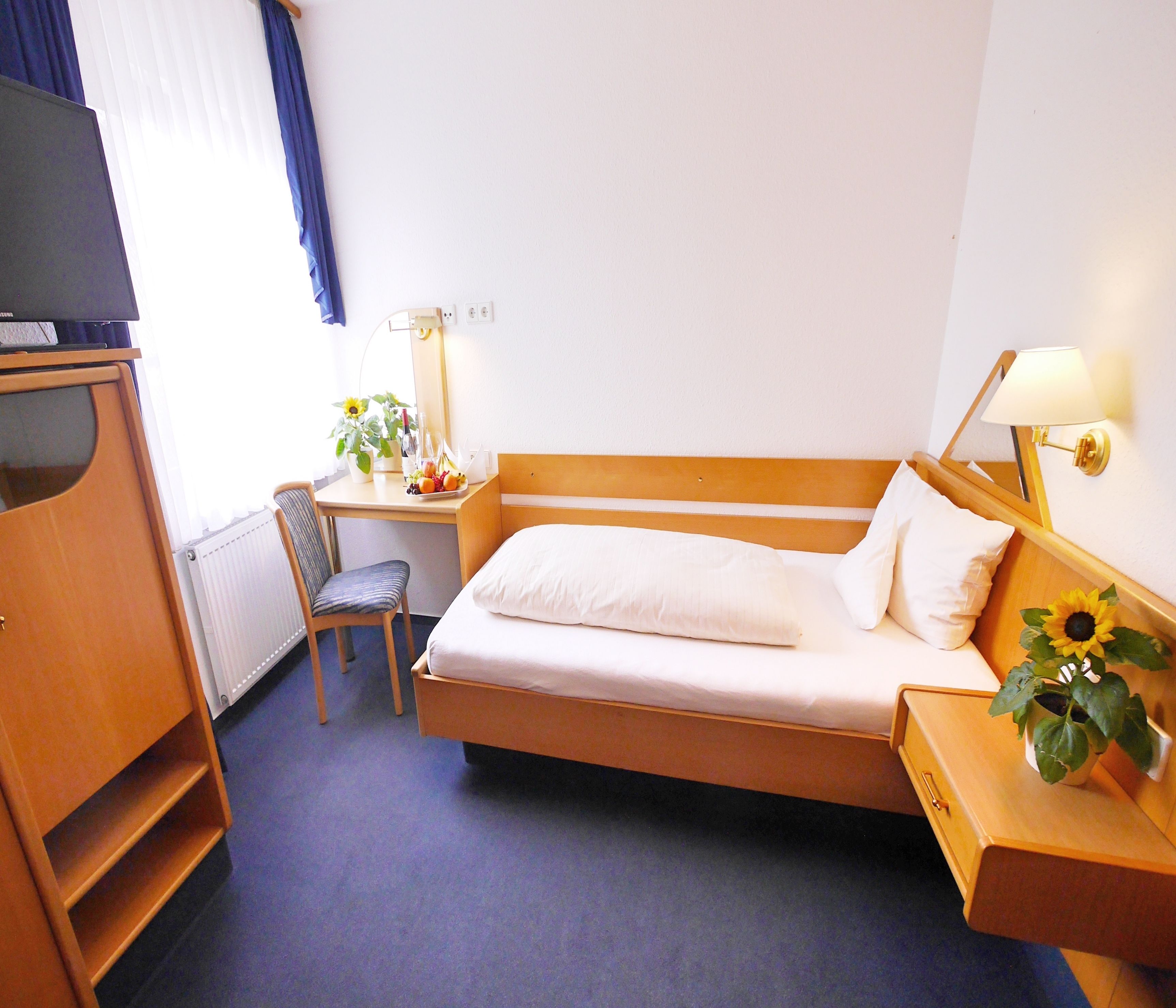 A hotel room with a single bed, a desk with a chair, a lamp, a radiator, and a television.